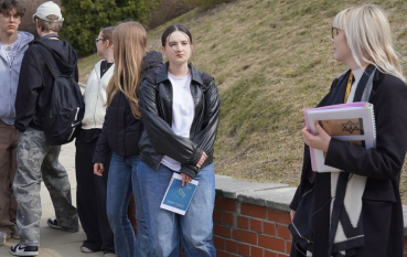 Dwie kobiety, jedna o jasnych blond włosach trzyma w rękach plik dokumentów, druga o ciemnych włosach trzyma w ręku niebieską mapę. 