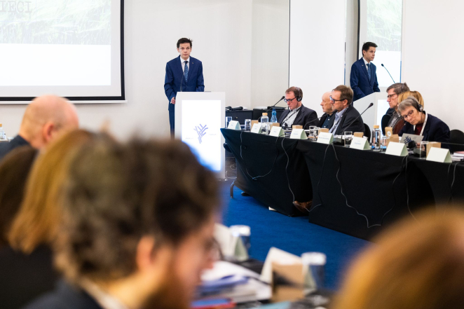 Mężczyzna przy mównicy dookoła ludzie w garniturach siedzących przy stołach. 