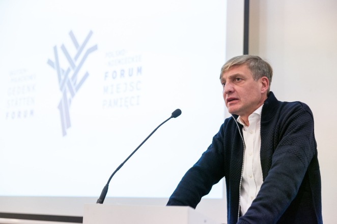 Starszy mężczyzna w białej koszuli i granatowym swetrze przemawia do mikrofonu.