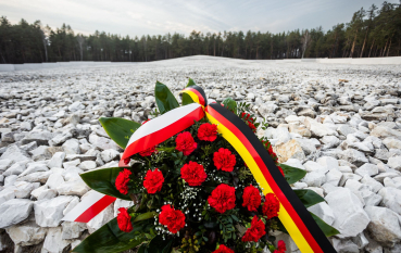 Wiązanka z czerwonych goździków opleciona wstęgą z flagą polską i niemiecką, w tle kopiec złożony z białych kamieni. 