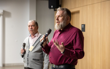 Dwóch mężczyzny, jeden z brodą przemawia do mikrofonu.