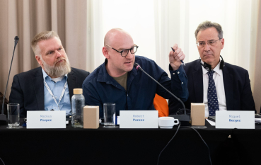 Trzech mężczyzn siedzi przy stole, łysy mężczyzna w okularach mówi do mikrofonu. 