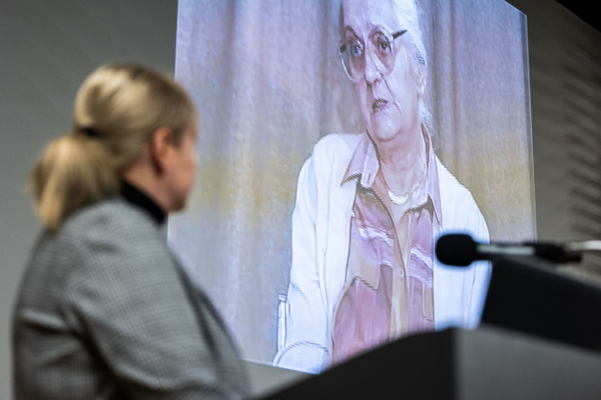 Osoba stojąca przy mównicy ogląda na dużym ekranie nagranie wideo przedstawiające starszą kobietę udzielającą świadectwa; scena odbywa się w przestrzeni konferencyjnej