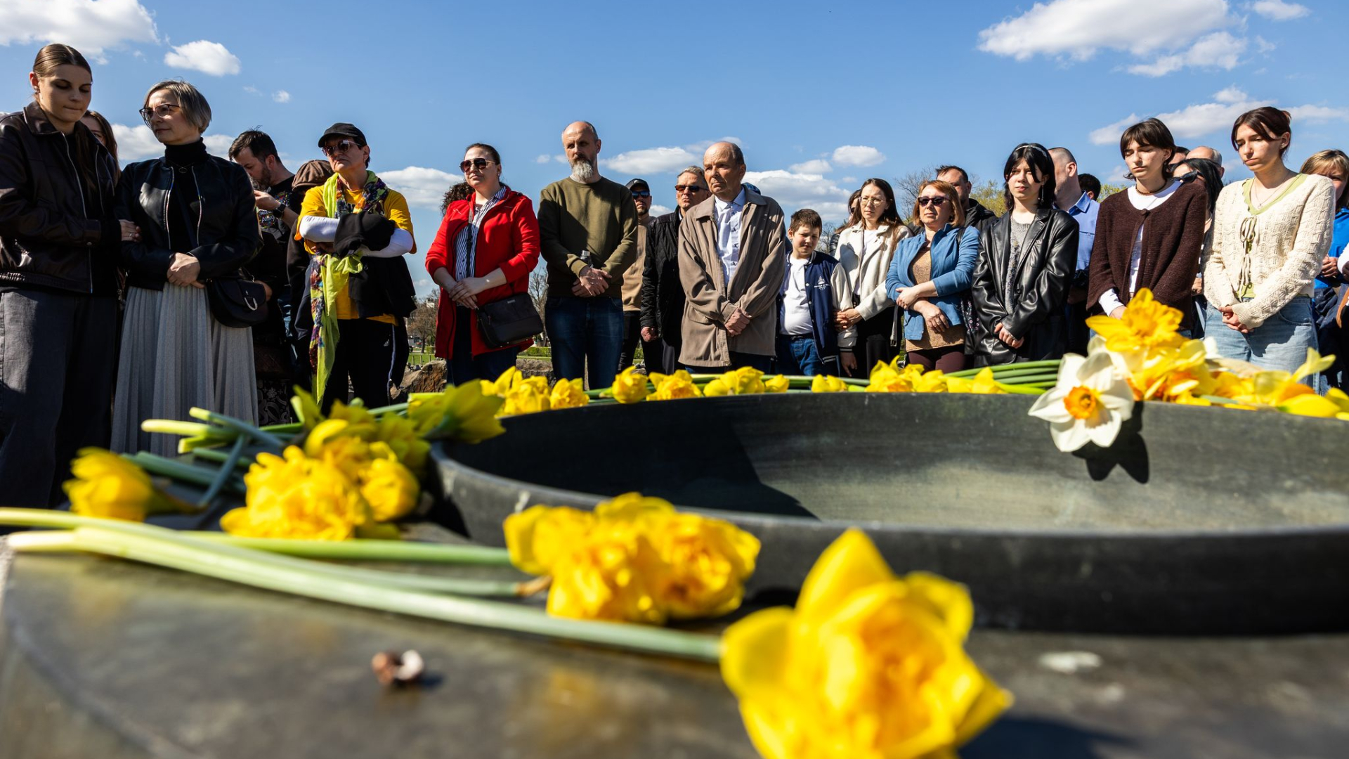 Grupa kilkudziesięciu osób, na pierwszym planie ułożona na postumencie żonkile.