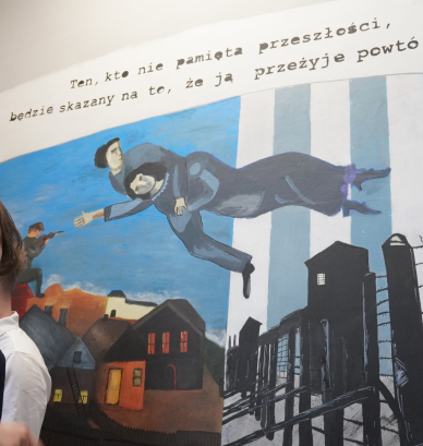 Uczniowie stoją przed muralem. Mural jest podzielony na dwie części. Po jego lewej stronie znajdują się namalowane domy, a na dachu jednego z nich siedzi skrzypek. Po prawej stronie muralu znajdują się druty kolczaste, a za nimi wieże strażnicze na tle biało-niebieskich pasów. Na środku muralu pomiędzy dwoma częściami unosi się postać mężczyzny, który trzyma kobietę. Nad postaciami widnieje napis: „Kto nie pamięta przeszłości, będzie skazany na to, że ją przeżyje powtórnie…”