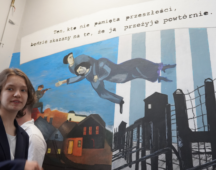 Uczniowie stoją przed muralem. Mural jest podzielony na dwie części. Po jego lewej stronie znajdują się namalowane domy, a na dachu jednego z nich siedzi skrzypek. Po prawej stronie muralu znajdują się druty kolczaste, a za nimi wieże strażnicze na tle biało-niebieskich pasów. Na środku muralu pomiędzy dwoma częściami unosi się postać mężczyzny, który trzyma kobietę. Nad postaciami widnieje napis: „Kto nie pamięta przeszłości, będzie skazany na to, że ją przeżyje powtórnie…”