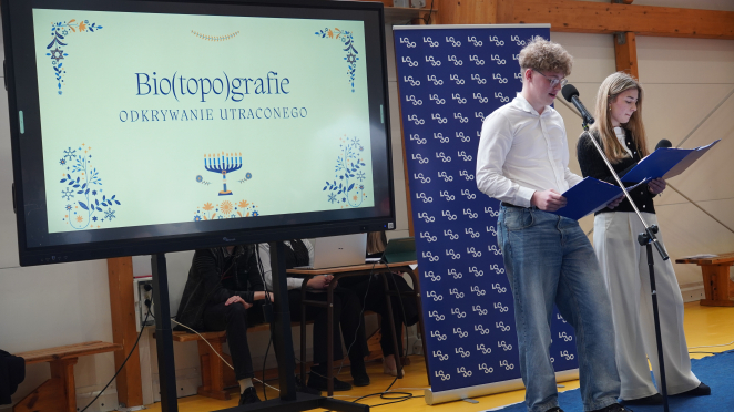 Uczeń i uczennica przemawiają do mikrofonu, trzymając podkładki z tekstem. Obok nich znajduje się ekran, na którym wyświetlana jest treść prezentacji multimedialnej. Tekst prezentacji to: „Bio(topo)grafie. Odkrywanie utraconego”.
