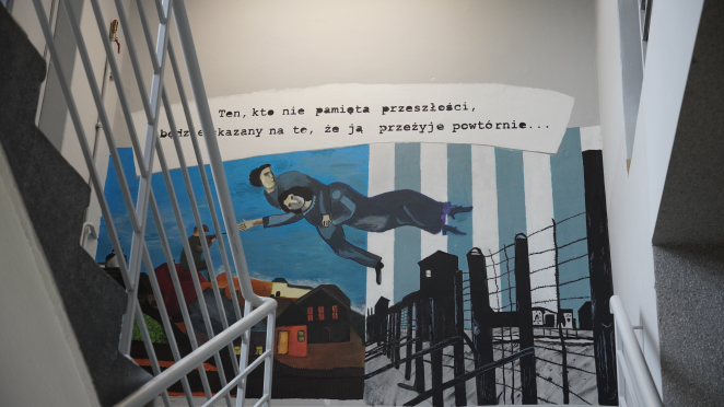 Mural: lewą część: domy oraz postać skrzypka na dachu zasłania balustrada i schody.; prawa część: druty kolczaste, wieże strażnicze na tle biało-niebieskich pasów; pośrodku: unosząca się postać mężczyzny i kobiety. Napis: „Kto nie pamięta przeszłości, będzie skazany na to, że ją przeżyje powtórnie…”