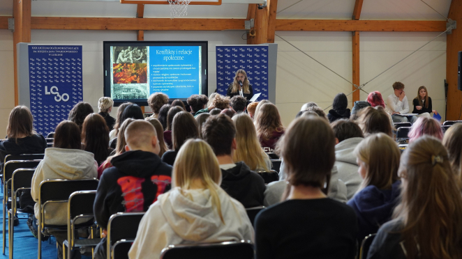 Grupa uczniów siedzi na krzesłach, przed nimi znajduje się ekran z prezentacją multimedialną. Na wyświetlonym slajdzie widnieje napis: „Konflikty i relacje społeczne”. Obok ekranu stoi przemawiająca uczennica.