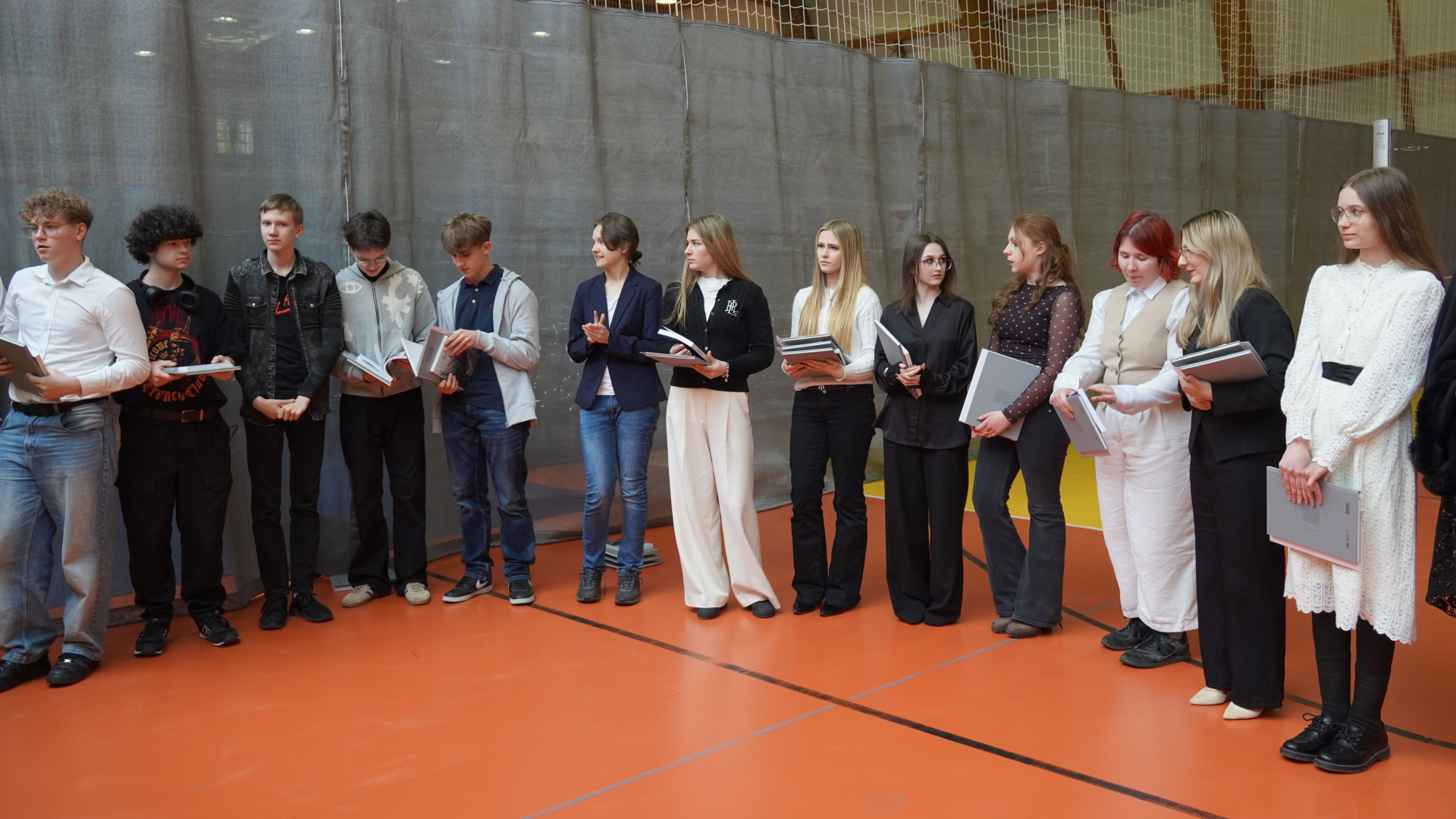 Uczniowie stoją w sali gimnastycznej. W rękach trzymają książki.