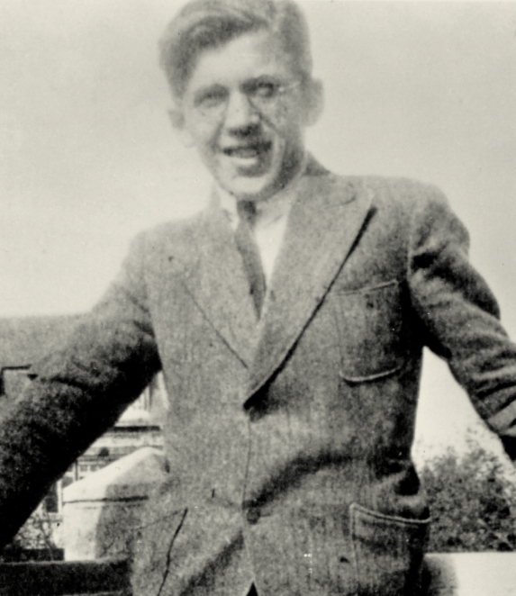 A black-and-white portrait photograph of a middle-aged man, taken from the chest upwards. His face is facing the camera; he has short, dark hair and is wearing a light-coloured shirt and a dark jacket. The background is plain and light in colour.