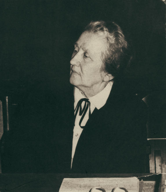 A black-and-white portrait photograph of a woman, taken from close up. Her face is turned slightly to one side; her hair is cropped short or tied back, and she is wearing dark clothing. The background is plain, with no visible details.