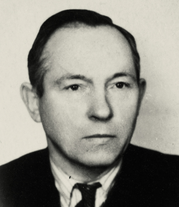 Black-and-white portrait photograph of a man, framed from the shoulders up. He has short hair and looks directly at the camera. The background is light.