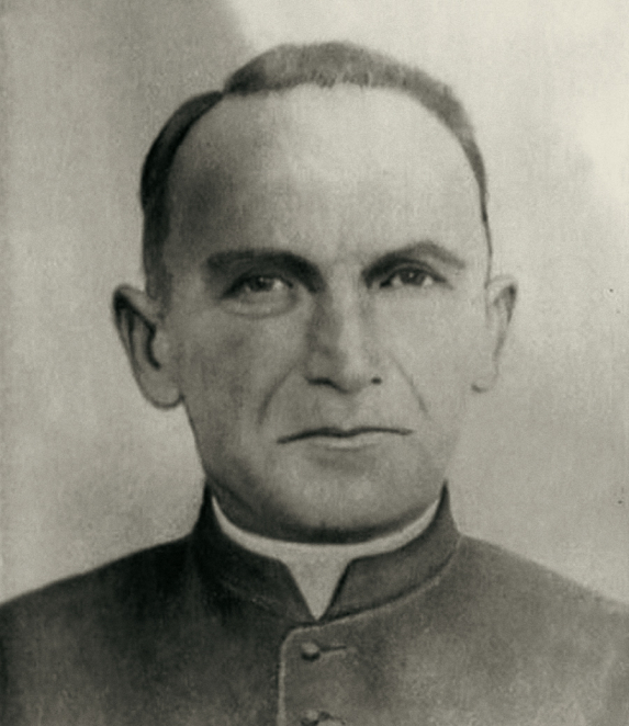 A black-and-white portrait photograph of a man, showing his head and shoulders. He has short hair and his face is turned slightly to one side. He is wearing a dark cassock with a white clerical collar. The background is light.