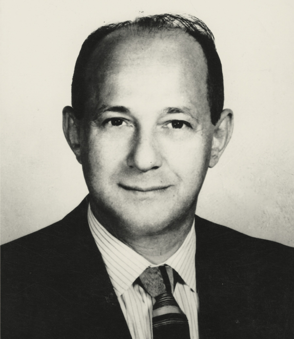 A black-and-white portrait photograph of a man, shot from the shoulders up. He has short hair and is facing forward. He is dressed in formal attire. The background is plain.