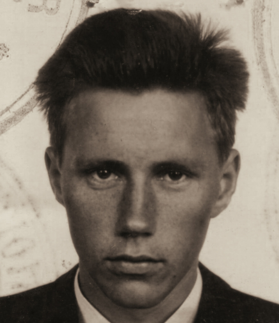 A black-and-white portrait photograph of a young man, showing his head and shoulders. He has short hair, is facing forward, and is wearing a light-coloured shirt. The background is plain.
