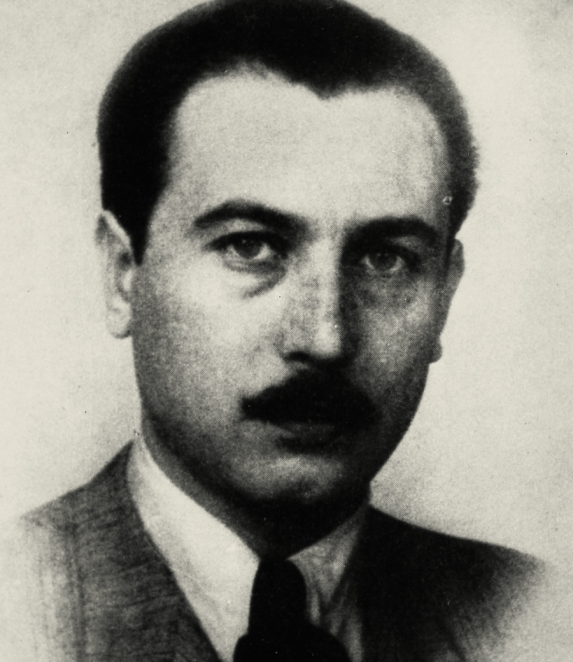 A black-and-white portrait photograph of a man, taken from close up. He is facing the camera, with short hair. He is wearing dark clothing. The background is light and lacks detail.