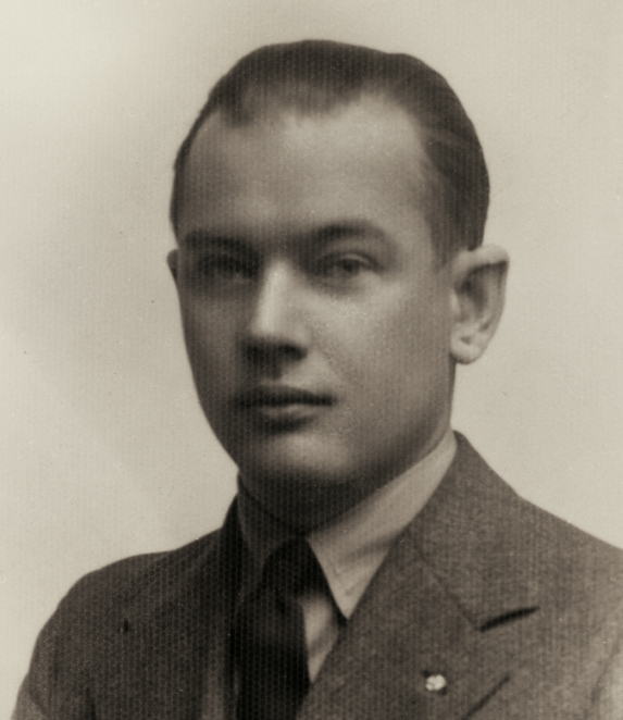 A black-and-white portrait photograph of a man, taken from the chest upwards. He has short hair and is facing the camera. He is wearing a jacket. The background is plain.