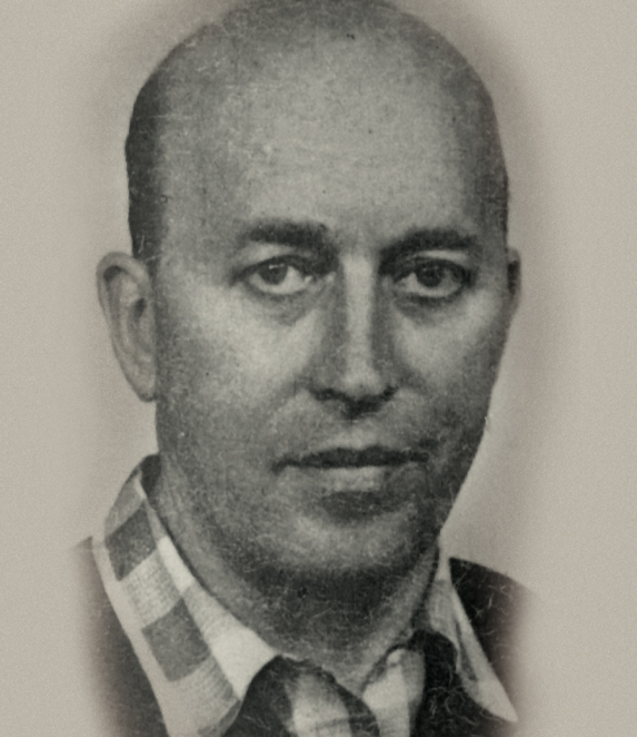 A black-and-white portrait photograph of a man, showing his head and shoulders. He has short hair and his face is turned slightly to one side. He is dressed in dark clothing. The background is plain.