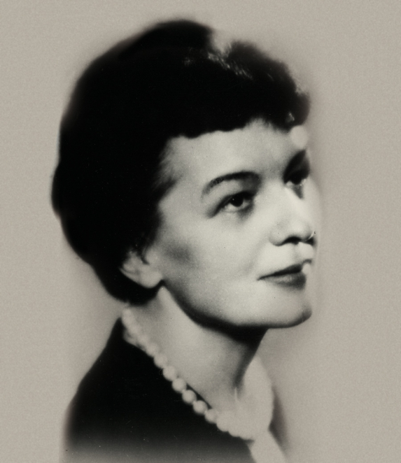 A black-and-white portrait photograph of a woman, taken from the chest upwards. Her hair is neatly styled, and her face is turned slightly to one side. She is dressed in dark clothing. The background is neutral.