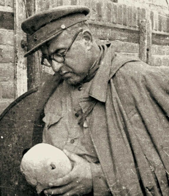 A black-and-white photograph of a middle-aged man wearing a military uniform. He is wearing a peaked cap and glasses. He is holding a human skull in his hands.