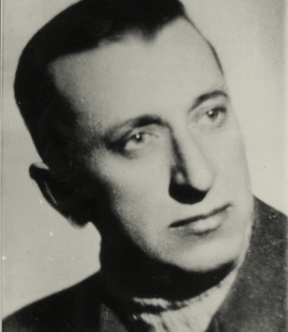 A black-and-white portrait photograph of a man, taken in close-up. He has short hair and is looking directly at the camera. He is wearing a jacket. The background is plain.