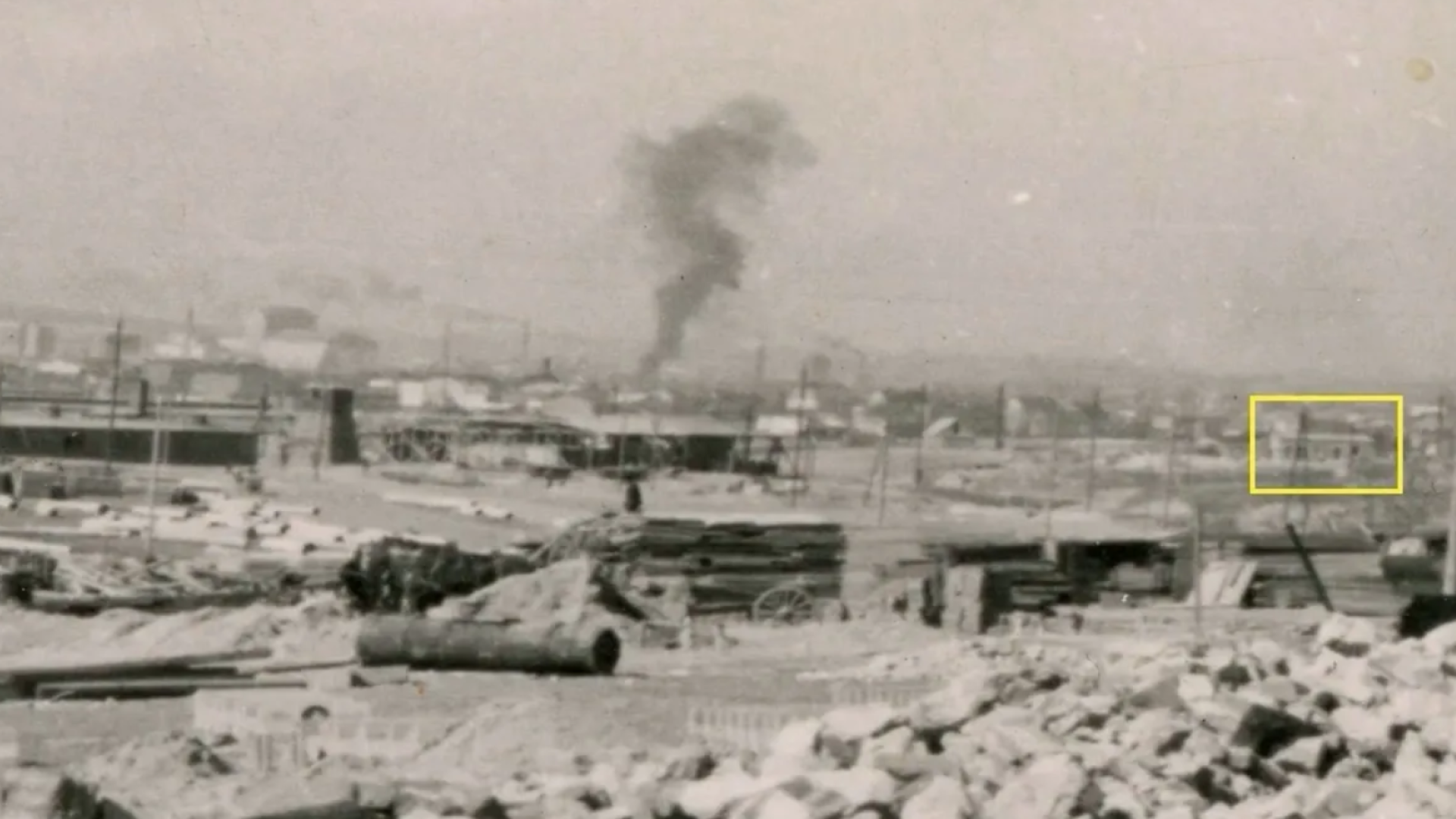 Black-and-white photo showing a destroyed area with buildings and smoke rising in the background. In the foreground, rubble and abandoned construction elements are scattered across the scene, while a small building on the right side is highlighted with a yellow frame.