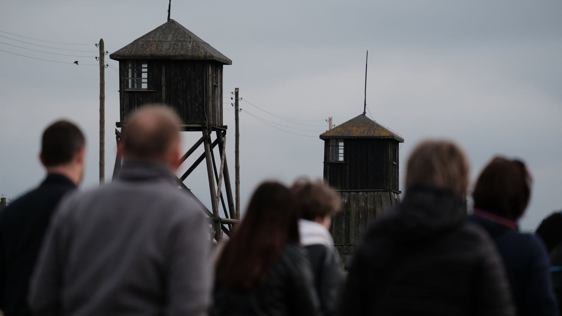 Zdjęcie przedstawia niewyraźnie zarysowaną na pierwszym planie grupę osób na otwartej przestrzeni. W tle, w oddali widoczne są drewniane wieże wartownicze charakterystyczne dla zabudowy obozu koncentracyjnego.