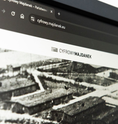 Zbliżenie na monitor komputera z otwartą stroną Cyfrowy Majdanek. Na ekranie widoczny jest adres serwisu oraz logo projektu, a poniżej archiwalne czarno-białe zdjęcie przedstawiające zabudowania obozowe.