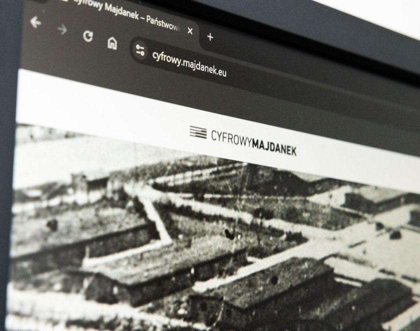 Zbliżenie na monitor komputera z otwartą stroną Cyfrowy Majdanek. Na ekranie widoczny jest adres serwisu oraz logo projektu, a poniżej archiwalne czarno-białe zdjęcie przedstawiające zabudowania obozowe.