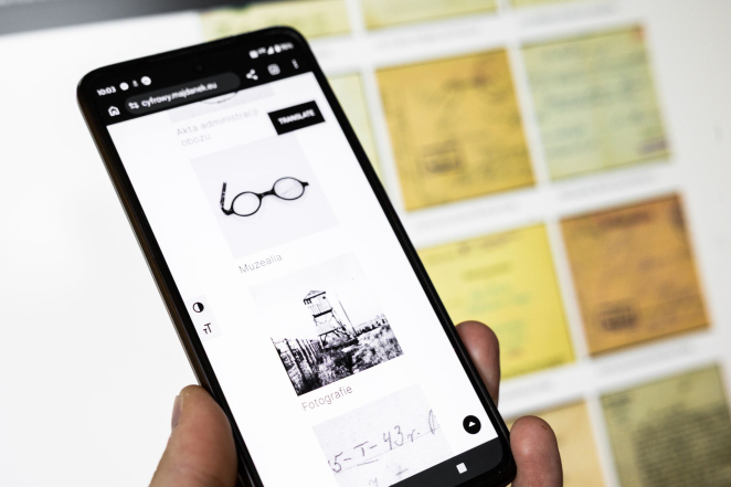 Dłoń trzymająca smartfon, na którego ekranie wyświetlane są zasoby cyfrowe serwisu Cyfrowy Majdanek. W tle, poza ostrością, widoczne są reprodukcje dokumentów archiwalnych.