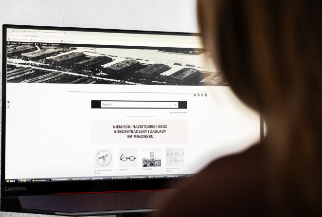 Zbliżenie na monitor komputera z otwartą stroną Cyfrowy Majdanek. Na ekranie widoczny jest adres serwisu oraz logo projektu, a poniżej archiwalne czarno-białe zdjęcie przedstawiające zabudowania obozowe.