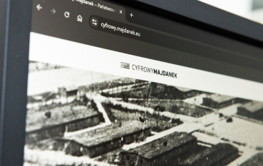 Zbliżenie na monitor komputera z otwartą stroną Cyfrowy Majdanek. Na ekranie widoczny jest adres serwisu oraz logo projektu, a poniżej archiwalne czarno-białe zdjęcie przedstawiające zabudowania obozowe.