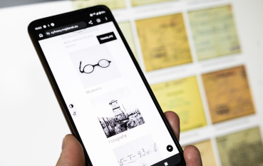 Dłoń trzymająca smartfon, na którego ekranie wyświetlane są zasoby cyfrowe serwisu Cyfrowy Majdanek. W tle, poza ostrością, widoczne są reprodukcje dokumentów archiwalnych.