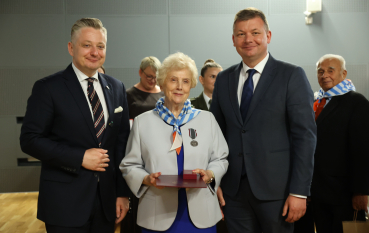 Dwóch mężczyzn w garniturach oraz kobieta z medalem. 