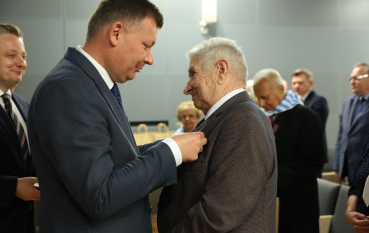 Mężczyzna w garniturze wpina medal w klapę marynarki starszego mężczyzny.