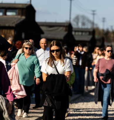 Grupa zwiedzających spaceruje po terenie byłego obozu koncentracyjnego na Majdanku. W tle widoczne są historyczne baraki obozowe, a osoby poruszają się alejką w słoneczny dzień.