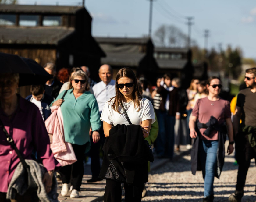 Grupa zwiedzających spaceruje po terenie byłego obozu koncentracyjnego na Majdanku. W tle widoczne są historyczne baraki obozowe, a osoby poruszają się alejką w słoneczny dzień.